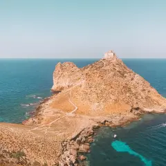Rocciosa isola con un antico fortino in cima, circondata da acque turchesi. Un sentiero serpeggia verso la vetta, evocando le spiagge delle isole Egadi.