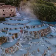 Cascate naturali con acqua azzurra e vapore, un casolare in pietra e due persone che si godono le terme di Saturnia.
