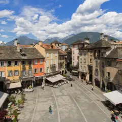 Piazza colorata con montagne sullo sfondo, un assaggio di dove si trova Domodossola.