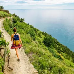 Donna con zaino cammina sul sentiero dell'Alta Via dei Monti Liguri, con il mare azzurro a destra e vigneti a sinistra.
