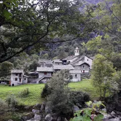 Borgo in pietra con campanile, immerso nel verde della Val Chiuse lla. Un ponte attraversa il torrente.