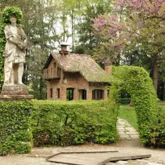 Un giardino incantato a Grazzano Visconti, dove si trova un'antica statua e una casetta in mattoni circondata da siepi verdi.