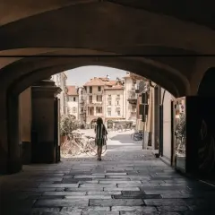 Una donna cammina sotto un portico verso una piazza soleggiata, con biciclette parcheggiate e palazzi storici che ricordano i paesi delle Langhe.