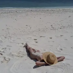 Person lying on a white sandy beach, wearing a straw hat, with the blue ocean in the background.
