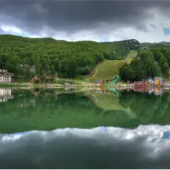 Lago alpino con riflessi di alberi e case, sullo sfondo il **passo del Cerreto** con piste da sci e giochi per bambini.