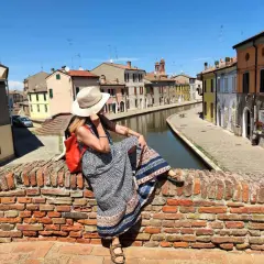Donna con cappello di paglia e zaino rosso seduta su un ponte di mattoni a Comacchio, con canali e case colorate. Un luogo ideale per una gita con bambini.