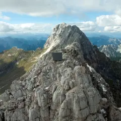 Bivacco Luca Vuerich, un cubo scuro su una cresta rocciosa, domina un panorama alpino mozzafiato sotto un cielo nuvoloso.