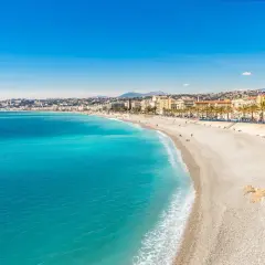 Spiaggia di ciottoli baciata dal sole sulla costa azzurra, con il mare turchese che lambisce la riva e palme che ombreggiano la Promenade des Anglais.