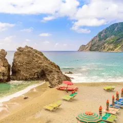 Spiaggia sabbia Liguria con scogli imponenti, mare azzurro, lettini e ombrelloni pronti per accogliere i bagnanti.