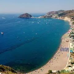 Vista aerea di una baia con spiaggia affollata, ombrelloni colorati e mare blu intenso. Un promontorio roccioso e un isolotto punteggiano l'orizzonte. Potrebbe essere una delle 5 spiagge più belle di Ischia.
