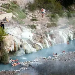 Persone si rilassano nelle acque fumanti delle terme di Petriolo, tra rocce calcaree e vegetazione.