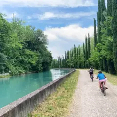 Bambini in bicicletta lungo la ciclabile Mincio, con un canale turchese e filari di cipressi.