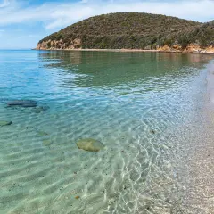 Acque cristalline e sabbia dorata nella Maremma Toscana. Il mare riflette il cielo azzurro, mentre la collina verdeggiante si affaccia sulla costa.