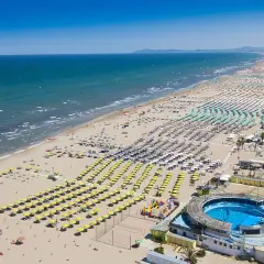 Vasta spiaggia della **costiera romagnola** con file ordinate di ombrelloni gialli e azzurri, mare calmo e un complesso con piscina.