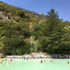 Piscina con persone che nuotano, circondata da vegetazione lussureggiante e rocce, un luogo che sembra uscito dalle grotte delle ninfe.