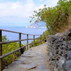 Sentiero in pietra con staccionata in legno, vista sul mare blu e una barca a vela. Perfetto per i cammini in Italia sul mare.