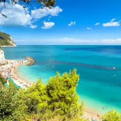 Spiaggia affollata con ombrelloni colorati e gente che nuota nelle acque cristalline. Una scogliera bianca domina la costa, incorniciata dal verde della vegetazione. Una perfetta località mare Marche.