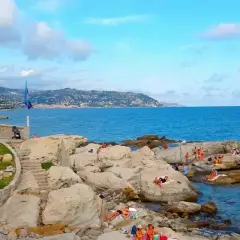 La chiesa bianca domina la costa rocciosa di Bordighera, con bagnanti che si godono il sole. Un luogo incantevole per scoprire cosa vedere a Bordighera.