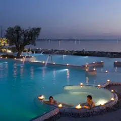 Coppia rilassata in una piscina termale illuminata da candele, con cascate e vista lago al tramonto. Un'oasi di pace alle terme a Brescia.