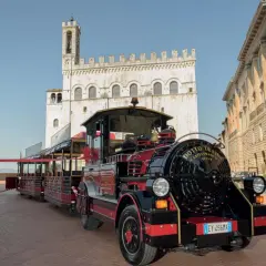 Il trenino turistico di Gubbio, rosso e nero, attende i passeggeri in una piazza storica con palazzi antichi.