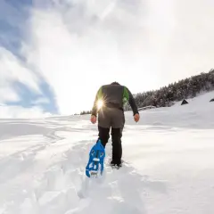 Escursionista con racchette da neve cammina su sentiero innevato, scoprendo posti belli in montagna Piemonte inverno. Sole splendente tra le nuvole.