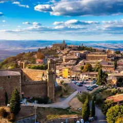 Scorci borghi antichi arroccati su una collina, con un castello imponente e case in pietra sotto un cielo azzurro con nuvole bianche.
