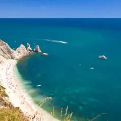 La spiaggia più bella del Conero, con sabbia bianca, mare cristallino e scogliere imponenti. Barche solcano le acque turchesi.