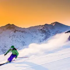 Due sciatrici sfrecciano su un pendio innevato al tramonto, con montagne maestose sullo sfondo. Un'emozione da provare per chi cerca dove sciare in Emilia Romagna.
