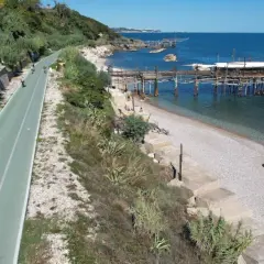 Ciclisti lungo la via verde della costa dei trabocchi, con il mare blu e le antiche macchine da pesca sullo sfondo.