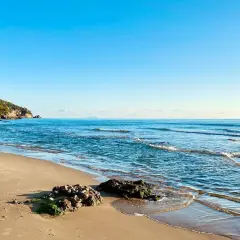 Spiaggia dorata e mare azzurro vicino a Roma. Le onde lambiscono rocce coperte di alghe, mentre una collina verde domina la costa.