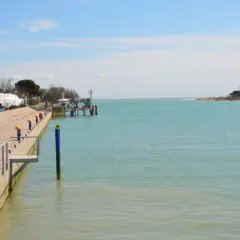 Un uomo guarda il mare calmo a Marano Lagunare, con barche coperte e un molo. La spiaggia lagunare è serena.
