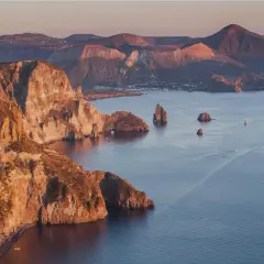 Scogliere dorate e mare blu intenso alle Isole Eolie, con piccole spiagge nascoste e faraglioni che emergono dall'acqua.