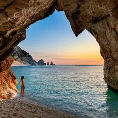 Vista da una grotta sulle spiagge della Riviera del Conero al tramonto. Una ragazza ammira il mare turchese e le rocce a picco.