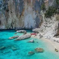 Spiaggia incantevole sulla costa di Baunei, con acque cristalline, rocce imponenti e una grotta marina. Kayak e persone godono del sole.