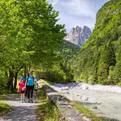 Famiglia in escursione ad Andalo, cammina lungo un sentiero con un fiume cristallino e montagne maestose sullo sfondo.