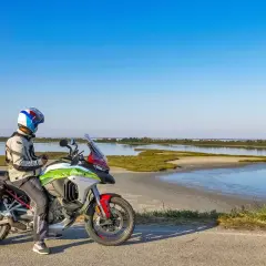 Motociclista con moto da enduro ferma sul ciglio di una strada sterrata, con sullo sfondo il paesaggio del Delta del Po.