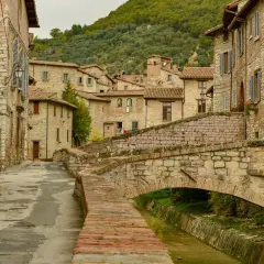 Antico ponte in pietra attraversa un ruscello, fiancheggiato da case in pietra e vicoli stretti, tipici dei paesi in Umbria.