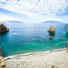 Spiaggia di ciottoli e acque cristalline, ideale per una vacanza alle Eolie. Barche a vela e isole all'orizzonte.