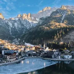 Villaggio alpino innevato ai piedi delle Dolomiti, ideale per escursioni invernali. Lago ghiacciato riflette le montagne.
