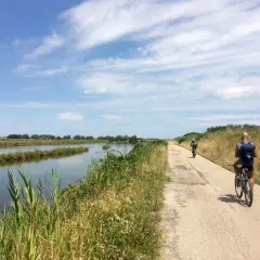 Cicloturismo in Emilia Romagna: due ciclisti pedalano su una strada sterrata lungo un canale, con vegetazione rigogliosa e cielo azzurro.
