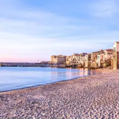La spiaggia di Cefalù al tramonto, con le case colorate che si affacciano sul mare calmo e la sabbia dorata.