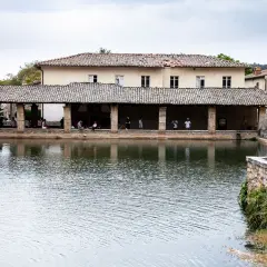 La piazza delle sorgenti termali di Bagni Vignone, con persone che si rilassano sotto un portico e il riflesso dell'acqua.