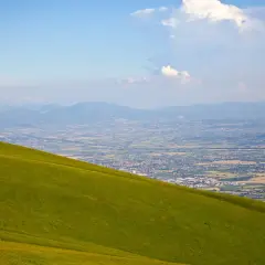 Prato verde scende dolcemente dal monte Subasio verso una valle punteggiata di case e montagne all'orizzonte.
