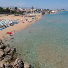 Una spiaggia affollata con lettini e ombrelloni, bagnanti in acqua e due kayak rossi sulla sabbia. Il noto mare cristallino invita a un tuffo.