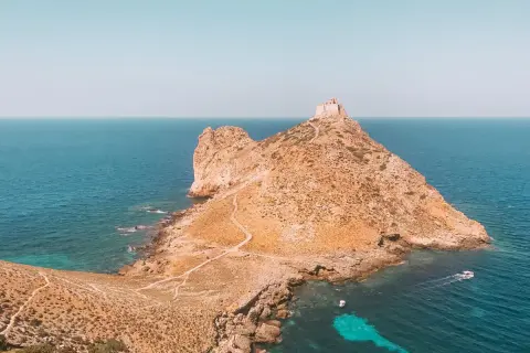 Rocciosa isola con un antico fortino in cima, circondata da acque turchesi. Un sentiero serpeggia verso la vetta, evocando le spiagge delle isole Egadi.