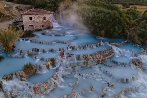 Cascate naturali con acqua azzurra e vapore, un casolare in pietra e due persone che si godono le terme di Saturnia.