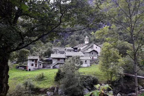Borgo in pietra con campanile, immerso nel verde della Val Chiuse lla. Un ponte attraversa il torrente.