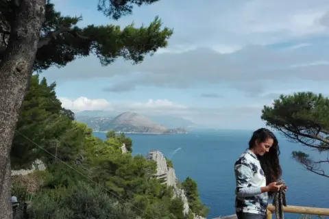 Donna con sciarpa guarda il cellulare, con vista mare e montagne di Capri a piedi.