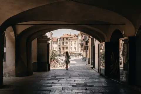 Una donna cammina sotto un portico verso una piazza soleggiata, con biciclette parcheggiate e palazzi storici che ricordano i paesi delle Langhe.