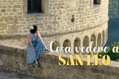 Donna seduta su un muretto di pietra a San Leo, con testo "Cosa vedere a San Leo".